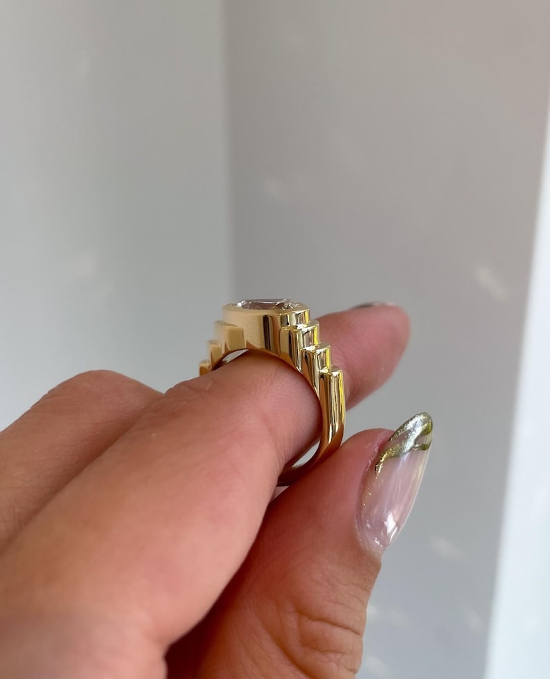 May include: A gold-colored ring with a rectangular, light-colored gemstone. The ring has a stepped design on the sides, creating a geometric pattern. The ring is held up by a person's fingers.