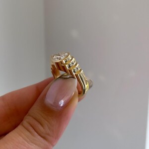 May include: A gold ring with a large, clear gemstone. The ring has a unique design with a series of stepped, textured elements on the band. The gemstone is oval-shaped and set in a bezel setting. The ring is held up by a person's hand.