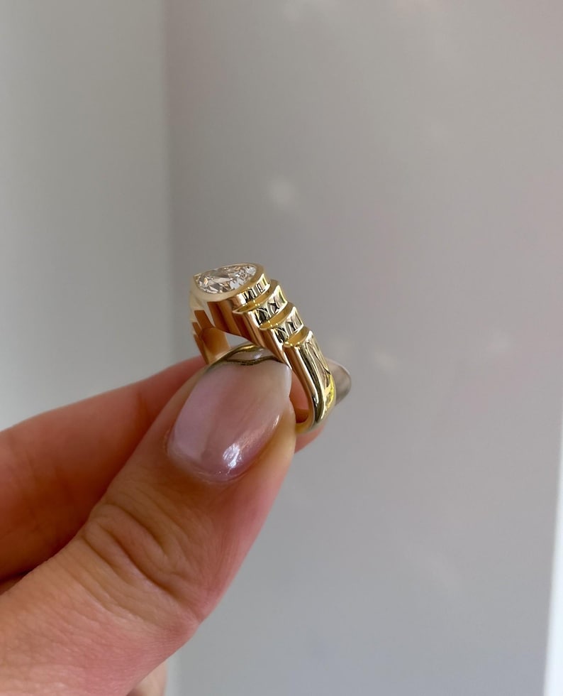 May include: A gold ring with a large, clear gemstone. The ring has a unique design with a series of stepped, textured elements on the band. The gemstone is oval-shaped and set in a bezel setting. The ring is held up by a person's hand.