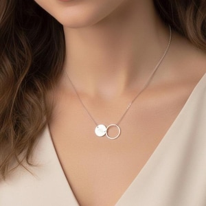 May include: A silver necklace featuring two connected pendants. One pendant is a small, engraved disc, and the other is a simple open circle. The necklace is worn against a neutral background, highlighting the jewelry's delicate design.