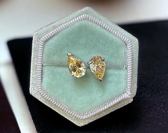 May include: Two pear-shaped yellow gemstones set in silver-toned metal prongs. The stones are positioned side-by-side, displaying a faceted cut that reflects light. The jewelry piece is displayed within a light green hexagonal velvet ring box.