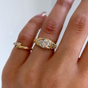 May include: Two gold rings. One ring features a pear-shaped diamond, set in a gold band with a textured design. The second ring is a simple gold band with small, clear stones. The rings are displayed on a hand.