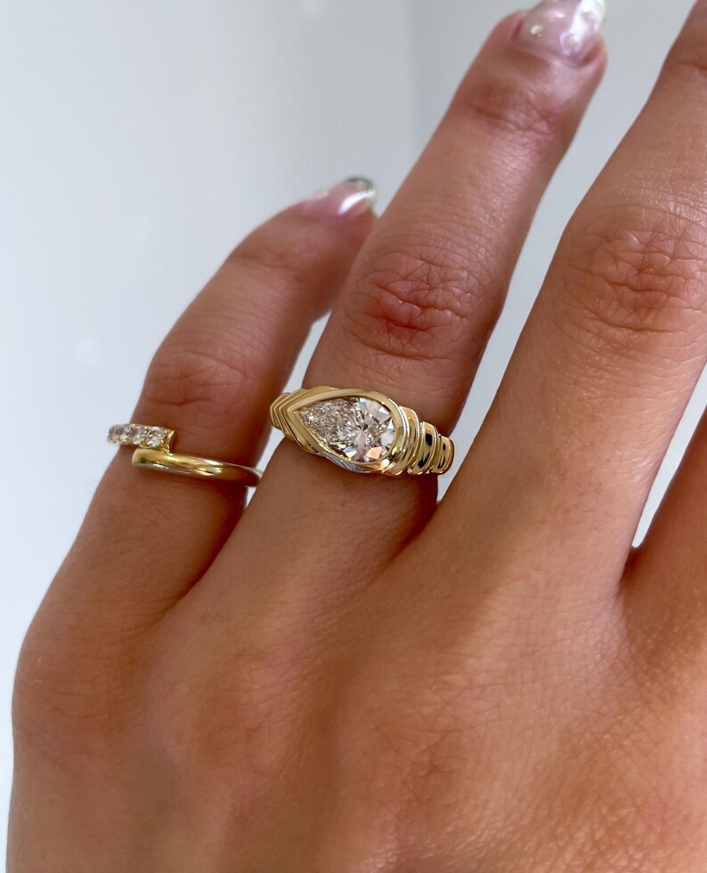 May include: Two gold rings. One ring features a pear-shaped diamond, set in a gold band with a textured design. The second ring is a simple gold band with small, clear stones. The rings are displayed on a hand.