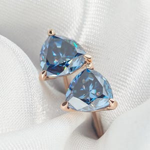 May include: A pair of stud earrings featuring trillion-cut blue gemstones set in rose gold metal. Each triangular stone is secured by three prongs, showcasing a faceted surface that reflects light with a clear, cool-toned blue hue.