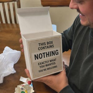 May include: A white rectangular gift box with an open lid. The front of the box features a beige label with black text that reads: THIS BOX CONTAINS NOTHING EXACTLY WHAT YOU WANTED. YOU'RE WELCOME. The box has a clean, minimalist design.