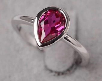 May include: A silver ring with a pear-shaped, bright pink gemstone. The gemstone is set in a bezel setting, and the band is polished silver. The ring is shown against a soft grey background. The gemstone is approximately 1.5 cm in length.