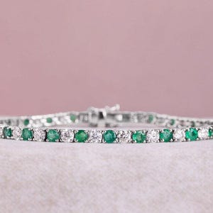 Puede incluir: Una pulsera de plata con piedras redondas alternas de esmeraldas y diamantes. La pulsera está sobre un fondo rosa claro, resaltando las piedras verdes y blancas. El cierre es visible, indicando un cierre seguro.