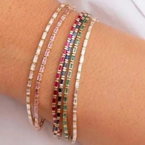 May include: Five delicate bracelets with rectangular gemstones in various colors: clear, pink, red, blue, and green. The bracelets are thin and appear to be made of gold, showcasing a minimalist design. The gemstones are set closely together.