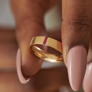 May include: A gold ring featuring a vertical, rectangular, ruby-coloured inlay. The ring is held between fingers with pink nail polish. The ring's band is a flat, polished gold. The ruby inlay provides a vibrant contrast against the gold.