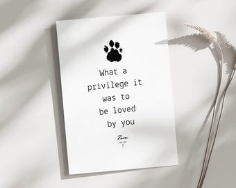 May include: A white rectangular print with a black paw print and the text "What a privilege it was to be loved by you." The print also includes a signature and a small heart symbol. A dried plant is to the right.