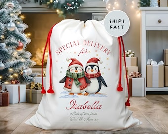 May include: A white Christmas gift sack with red drawstring closure. The sack features a watercolor illustration of two penguins wearing winter attire. Text on the sack reads "Special Delivery for Isabella" and "Lots of love from Dad & Mom xo."
