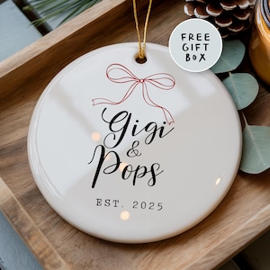 May include: A white ceramic ornament with a red bow design and the words "Gigi & Pops" in elegant script. The year "EST. 2025" is printed below. A gold string is attached for hanging. A small circle reads "FREE GIFT BOX".