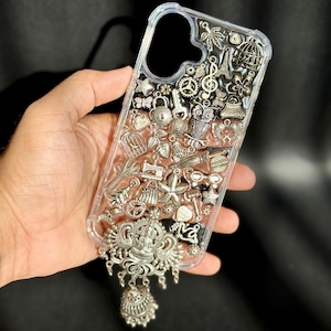 Puede incluir: Funda transparente para teléfono con abalorios plateados, como una jaula para pájaros, la Torre Eiffel y engranajes. La funda tiene un dije plateado colgante con una figura detallada y un colgante en forma de campana. La funda se sostiene en una mano.