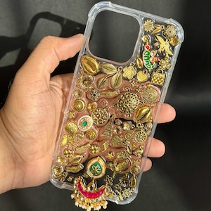 Op de afbeelding: Transparante telefoonhoes versierd met een reeks goudkleurige bedels en versieringen. De hoes heeft verschillende vormen, waaronder bloemen, bladeren en geometrische ontwerpen, met een roze-bruine achtergrond. Een decoratieve, rood-witte hanger hangt aan de onderkant.