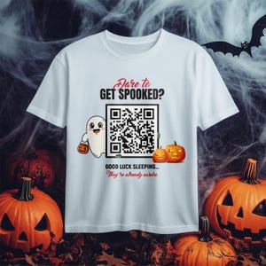 Könnte beinhalten: Weißes T-Shirt mit Halloween-Motiv. Das Shirt zeigt einen Geist mit einem Kürbis, zwei orangefarbene Kürbisse und den Text "Dare to Get Spooked?" und "Good Luck Sleeping... They're already awake."