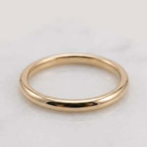 Op de afbeelding: Een gepolijste gouden trouwring. De ring heeft een glad, afgerond profiel dat licht reflecteert. Deze klassieke band is geschikt voor dagelijks gebruik. De ring wordt gepresenteerd op een wit marmeren oppervlak.