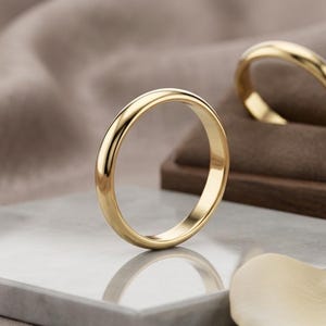 May include: Two gold wedding bands. One ring stands upright on a gray marble surface, reflecting light. The other ring rests on a brown velvet cushion inside a wooden box. A cream-colored petal is in the foreground.