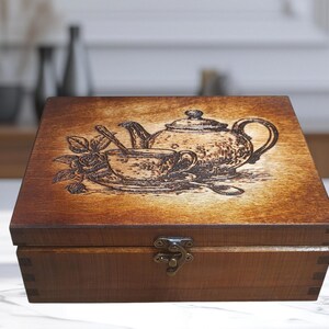 May include: A wooden tea box with a hinged lid and a metal clasp. The lid features a detailed etched design of a teapot, teacup, spoon, and rose. The box is stained in a warm brown color.