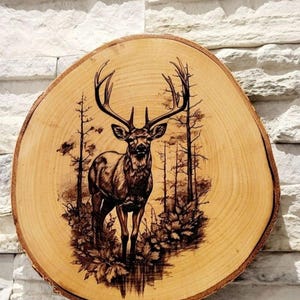Könnte beinhalten: Runde Holzplakette mit einer Hirschszene. Der Hirsch steht im Mittelpunkt, umgeben von Bäumen und Blättern. Das Kunstwerk ist in das Holz eingebrannt, was einen rustikalen, natürlichen Look erzeugt. Die Plakette hat einen Durchmesser von etwa 30 cm.