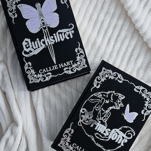 Op de afbeelding: Twee zwarte hardcover boeken met witte en zilveren decoratieve details. Eén boek heeft een vlinder en de titel "Quicksilver". Het andere boek heeft een wolf illustratie en de titel "Brimstone". Beide boeken zijn van Callie Hart.