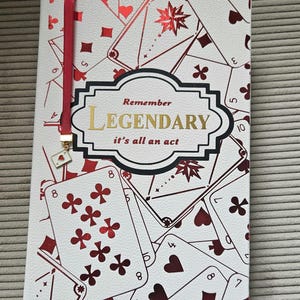 Puede incluir: Diario blanco con diseño de naipes y marcapáginas de cinta roja. La portada presenta palos de naipes y las palabras "Remember Legendary it's all an act" en oro y negro.