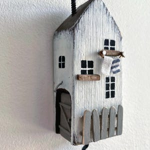 Peut inclure: Un crochet mural décoratif en forme de maison en bois peint en blanc avec des détails noirs. Il présente une cheminée noire, des fenêtres, une petite clôture et un crochet. Une petite serviette à rayures bleues est suspendue. Thème nautique rustique.