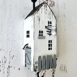 Op de afbeelding: Een witte, houten wandhaak in de vorm van een huis met een zwarte schoorsteen, ramen en een klein hekje. Het huis heeft een verweerde, rustieke uitstraling. Onder het huis hangt een zwarte haak.