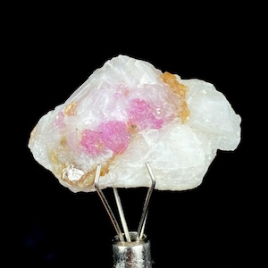 Może przedstawiać: Surowy, biały okaz minerału z plamami jasnoróżowego i brązowego koloru. Kamień jest trzymany przez srebrny metalowy zacisk na czarnym tle. Różowe obszary wydają się krystaliczne.