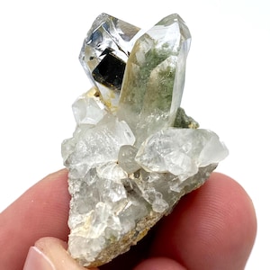 Puede incluir: Un grupo de cristales de cuarzo transparente con una inclusión verde similar al musgo. Los cristales son multifacéticos y se asientan sobre una matriz rugosa de color claro. Los cristales están sostenidos por los dedos de una persona.