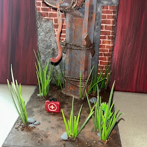 May include: A miniature diorama featuring a weathered, rust-colored metal I-beam structure against a brick wall backdrop. A curved metal hook hangs from the beam, secured with twine. The base includes green plastic grass, small gray stones, and a red first aid kit.