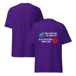 May include: Purple t-shirt with the text "DEL DICHO AL HECHO HAY MUCHO TRECHO". The design includes a teal frog and a red hibiscus flower. The back of the shirt is plain purple.