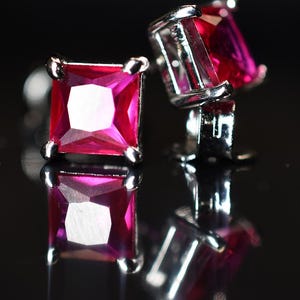 Peut inclure: Gros plan de deux paires de boucles d'oreilles à clous en forme de carré, avec des pierres précieuses rouge rubis. Les boucles d'oreilles sont serties dans un métal argenté et se reflètent sur une surface noire brillante.