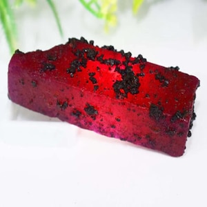 Puede incluir: Un objeto rectangular rojo vibrante con manchas oscuras y texturizadas. El artículo tiene un aspecto ligeramente brillante y está sobre un fondo blanco. La impresión general es la de un alimento, posiblemente un postre o un tipo de caramelo.