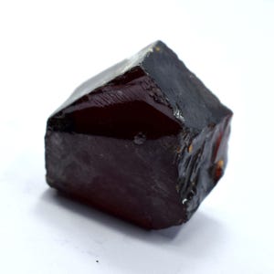 May include: A dark red, faceted gemstone with a rough, natural texture. The gem has a deep, translucent color and a slightly irregular shape, suggesting it is a raw, uncut stone. The stone is set against a white background.