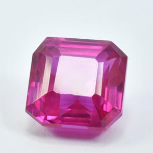 May include: A faceted, rectangular-cut, hot pink gemstone. The gem has a clear, reflective center and multiple facets that catch the light. The gemstone is set against a white background.
