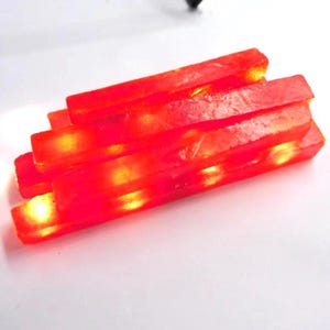 Op de afbeelding: Een stapel rechthoekige, doorschijnende, rode staven met een gloeiend, geel-oranje interieur. De staven zijn op elkaar gestapeld, waardoor een gelaagd effect ontstaat. De algehele uitstraling suggereert een decoratief of artistiek element.