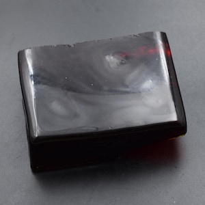 May include: A rectangular soap bar in a dark amber hue, with a glossy surface. The top edge of the soap is slightly uneven, while the rest of the surface is smooth and polished. The colour is a deep, translucent brown.