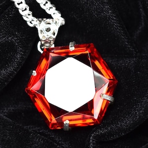 Può includere: Collana d'argento con un pendente esagonale rosso acceso. Il pendente ha un centro bianco e dettagli argentati. La catena è a maglie classiche, su uno sfondo di tessuto scuro e strutturato.