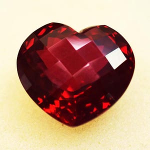 May include: A faceted, heart-shaped gemstone in a deep red hue. The gem's surface reflects light, showcasing its multiple facets. The heart shape is clearly defined, with a polished appearance. The gemstone is set against a light yellow background.