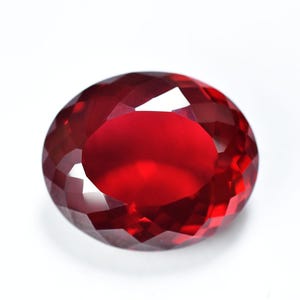 Op de afbeelding: Een gefacetteerde, ovaalvormige, dieprode edelsteen. De edelsteen heeft een briljant, reflecterend oppervlak met meerdere facetten die het licht vangen en weerkaatsen. De steen is geplaatst tegen een witte achtergrond.