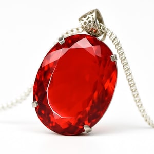 Peut inclure: Un pendentif en forme d'ovale avec une pierre précieuse rouge. La pierre rouge est facettée et transparente. Le pendentif est suspendu à une chaîne argentée. Le fermoir est orné de motifs décoratifs.