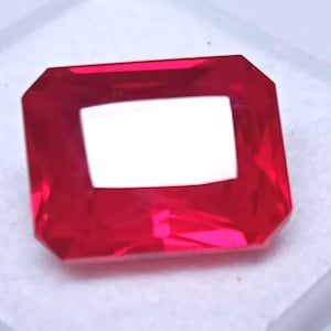 May include: A faceted, rectangular-cut, red gemstone with a bright, reflective surface. The gem has a large, white, rectangular opening in the center. The gemstone is set against a white background.