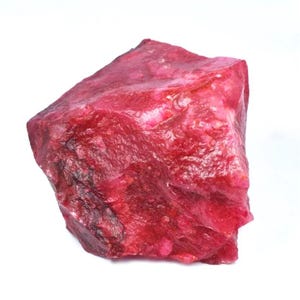Peut inclure: Une pierre précieuse de rubis brute, de couleur rouge intense et profonde. La surface est texturée et irrégulière. Le rubis est présenté sur un fond blanc uni.