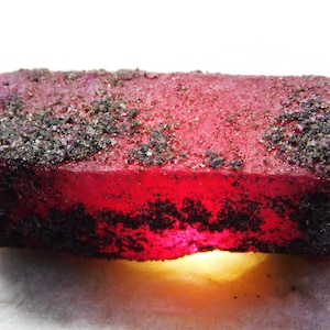 May include: A rectangular, raw gemstone with a deep red hue, accented by dark, granular inclusions on the surface. The stone has a rough texture and a glowing, amber-colored light emanating from its base.