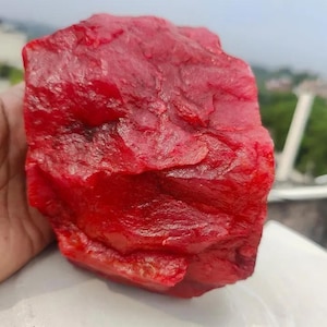 May include: A large, roughly textured, deep red gemstone. The stone has an irregular shape with a slightly glossy surface. The colour is a vibrant, saturated red, with variations in tone across the surface.