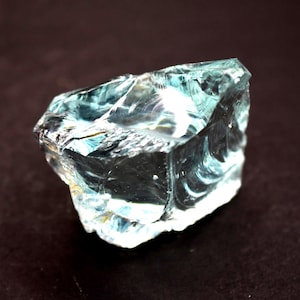 May include: A rough-cut, translucent blue-green gemstone with a crystalline structure. The stone has a multifaceted surface, reflecting light and creating a shimmering effect. The gemstone is set against a dark background.