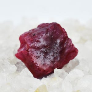 May include: A raw, uncut ruby gemstone with a deep red hue. The rough stone has an irregular shape and a textured surface. It is set against a backdrop of small, clear crystals, enhancing its color and natural beauty. This gemstone is ideal for jewelry making or collecting.