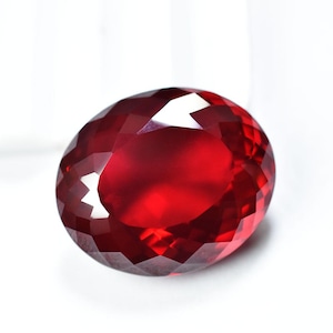 Puede incluir: Una gema facetada, de forma ovalada, de color rojo intenso. La gema tiene un corte brillante con numerosas superficies reflectantes, creando un efecto brillante. La piedra se encuentra sobre un fondo blanco liso, resaltando su color y claridad.