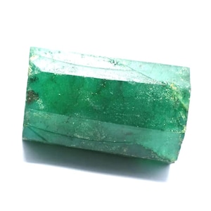 May include: A rectangular, raw emerald gemstone with a deep green hue. The gem has a faceted surface with visible inclusions and imperfections. The edges are rough and uneven, showcasing its natural state.