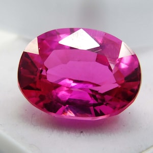 Può includere: Una gemma sfaccettata di forma ovale in un vibrante colore magenta. La superficie della gemma riflette la luce, mostrando le sue molteplici sfaccettature. La gemma è posta su uno sfondo bianco, evidenziando il suo colore e la sua chiarezza. Per gioielli.